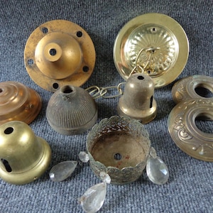 May include: A collection of vintage brass lighting components including ceiling canopies, socket covers, and a decorative metal bobeche with three hanging clear glass crystals. The set features various metallic finishes and intricate embossed patterns.