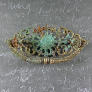 May include: An oval-shaped metal drawer pull featuring an intricate openwork design with a central floral motif. The hardware displays a weathered brass finish with prominent teal-colored oxidation throughout the ornate scrollwork and handle.