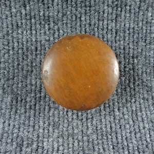 May include: A circular, smooth, brown wooden knob with a polished finish. The object features a slightly domed top surface with natural wood grain patterns and minor surface variations, presenting a simple, functional, and rounded hardware design.