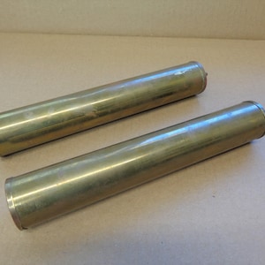 May include: Two cylindrical brass tubes with rounded ends. The tubes have a polished, reflective surface, and are lying parallel to each other. The brass has a warm, golden hue, and the tubes appear to be of similar size.