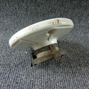 May include: A white ceramic ceiling light fixture base with a circular, slightly domed shape. The piece features a metallic mounting bracket attached to the underside, showing signs of oxidation and wear along the edges of the ceramic surface.