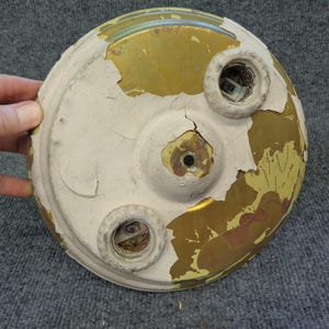 May include: A round, aged, off-white and gold-colored object with three circular openings. The paint is peeling, revealing the gold layer beneath. The object is held by a hand, suggesting its size and scale.