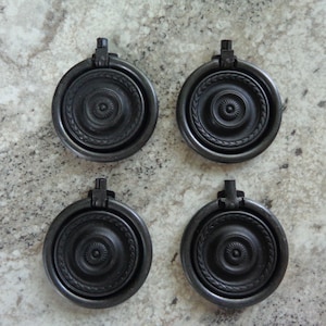May include: Four black metal ring pulls with a decorative design. The pulls have a circular shape with a raised center and a ring on the top.