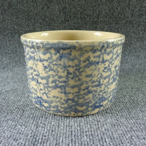 May include: A round, ceramic crock with a cream-colored base and a blue speckled pattern. The crock has a slightly flared rim and a smooth, unglazed interior. The exterior is decorated with a mottled blue design, giving it a rustic, vintage appearance.