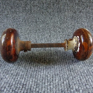 May include: A pair of round, brown, mottled ceramic door knobs connected by a central metal spindle. The knobs feature a glossy, marbled finish with dark and light brown tones, and the metal components show signs of oxidation and wear.