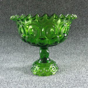 May include: A green glass pedestal bowl with a scalloped rim. The bowl features a pattern of circles and ovals. The stem and base are also decorated with circular designs. The bowl is approximately 7 inches tall.