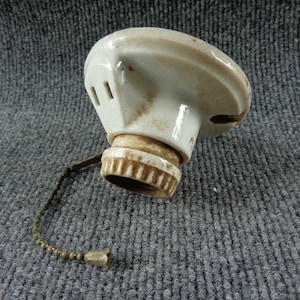 May include: A white ceramic light socket fixture featuring a circular mounting base, a threaded bulb receptacle, and a metallic pull chain with a small bell-shaped end piece. The ceramic surface shows signs of age with brown discoloration.