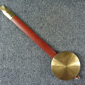 May include: A clock pendulum featuring a long, flat, reddish-brown wooden rod attached to a polished brass top connector and a circular, slightly conical brass bob at the bottom, showing a metallic sheen and subtle surface texture.