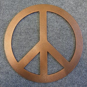 May include: A copper-colored peace sign with a textured finish. The symbol is a circle with a vertical line and two diagonal lines inside. The peace sign is displayed on a gray, textured surface.