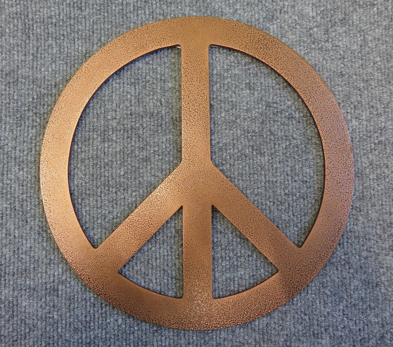 May include: A copper-colored peace sign with a textured finish. The symbol is a circle with a vertical line and two diagonal lines inside. The peace sign is displayed on a gray, textured surface.