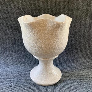 May include: A cream-colored pedestal bowl featuring a textured, pebbled exterior surface and a scalloped, wavy rim. The vessel stands on a circular base that tapers into a narrow stem, supporting the wide, rounded bowl with a smooth interior finish.