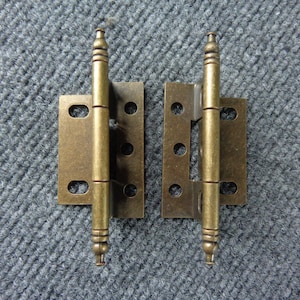 May include: Two antique brass-colored cabinet hinges. Each hinge has a rectangular base with multiple screw holes and a decorative, vertical barrel. The hinges are designed for furniture or cabinet doors.