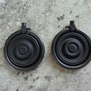 May include: Two black metal ring pulls with a decorative design. The pulls have a raised center with a circular pattern and a ring around the outside.
