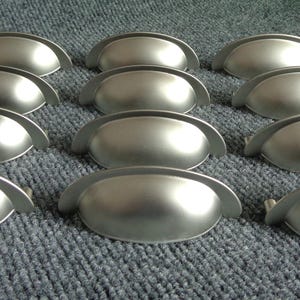 May include: A set of 12 silver cabinet knobs with a rounded, slightly curved shape. The knobs are arranged in rows on a gray surface.