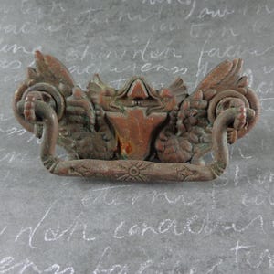 May include: A detailed, antique bronze drawer pull with ornate, winged details and a central shield design. The pull features two curved handles and measures approximately 5 inches in width.