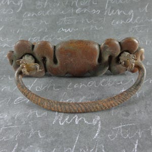 May include: A vintage-style, brown metal drawer pull with a decorative, ornate design. The pull features a curved handle with a textured pattern and a central, rounded section. The hardware is likely for a cabinet or drawer.