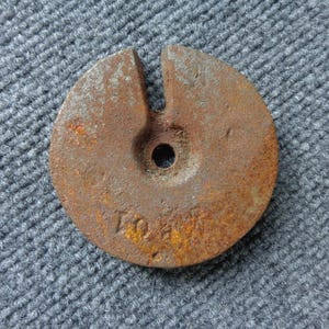 May include: A rusty, circular metal weight with a slot cut into the top and a central hole. The weight has the number "10" stamped on its surface. The object is set against a textured, gray background.