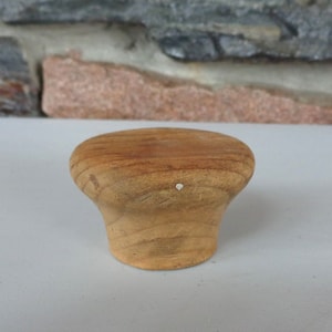May include: A wooden knob with a rounded top and a slightly flared base. The knob is a light brown color and has a small white mark on the top.