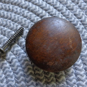 May include: A round, dark brown wooden cabinet knob with a smooth, worn finish and a visible grain pattern. Beside the knob lies a single silver-colored metal screw with a threaded shank, designed for mounting the knob to a drawer or door surface.