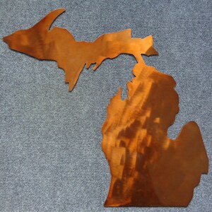 May include: A flat, copper-colored metal wall art piece shaped like the state of Michigan, including both the Upper and Lower Peninsulas. The surface features a polished, reflective finish with visible brushed metal textures and warm brown tones.