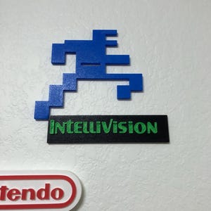May include: A blue pixelated figure, similar to the character from the video game 'Space Invaders', is mounted on a white wall. Below the figure is a black rectangular sign with the text "Intellivision" in green letters.