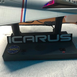 Może przedstawiać: Model statku kosmicznego w kolorze srebrnym z czerwonymi i niebieskimi paskami. Na podstawie napis "ICARUS", logo NASA i przyciski. Tylna część statku kosmicznego świeci.