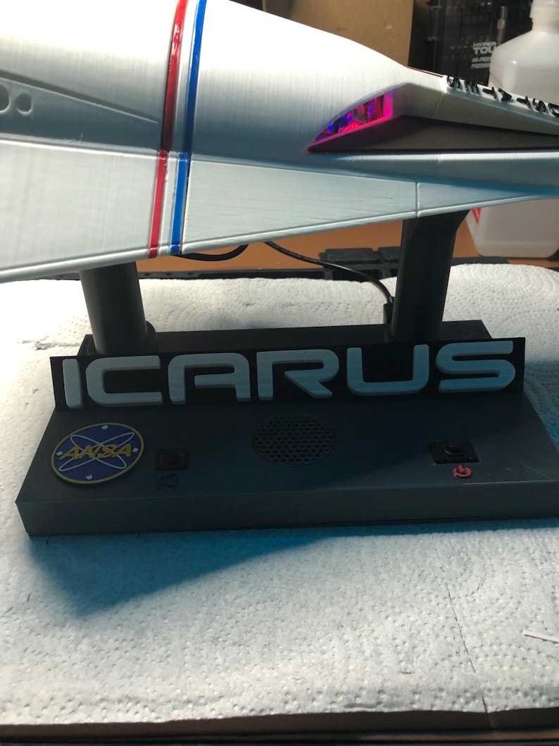 Może przedstawiać: Model statku kosmicznego w kolorze srebrnym z czerwonymi i niebieskimi paskami. Na podstawie napis "ICARUS", logo NASA i przyciski. Tylna część statku kosmicznego świeci.