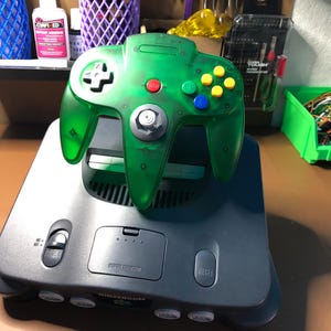 May include: A green translucent Nintendo 64 controller with yellow, blue, and red buttons sits atop a gray Nintendo 64 console. The console has a power button, reset button, and a memory expansion slot.