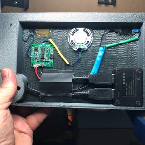 Może przedstawiać: Zbliżenie wnętrza czarnego urządzenia elektronicznego. Widoczne elementy to mały głośnik, zielona płytka drukowana oraz r&oacute;żne przewody i złącza. Urządzenie posiada r&oacute;wnież dwa porty USB i czarną skrzynkę z napisem "CKCS".