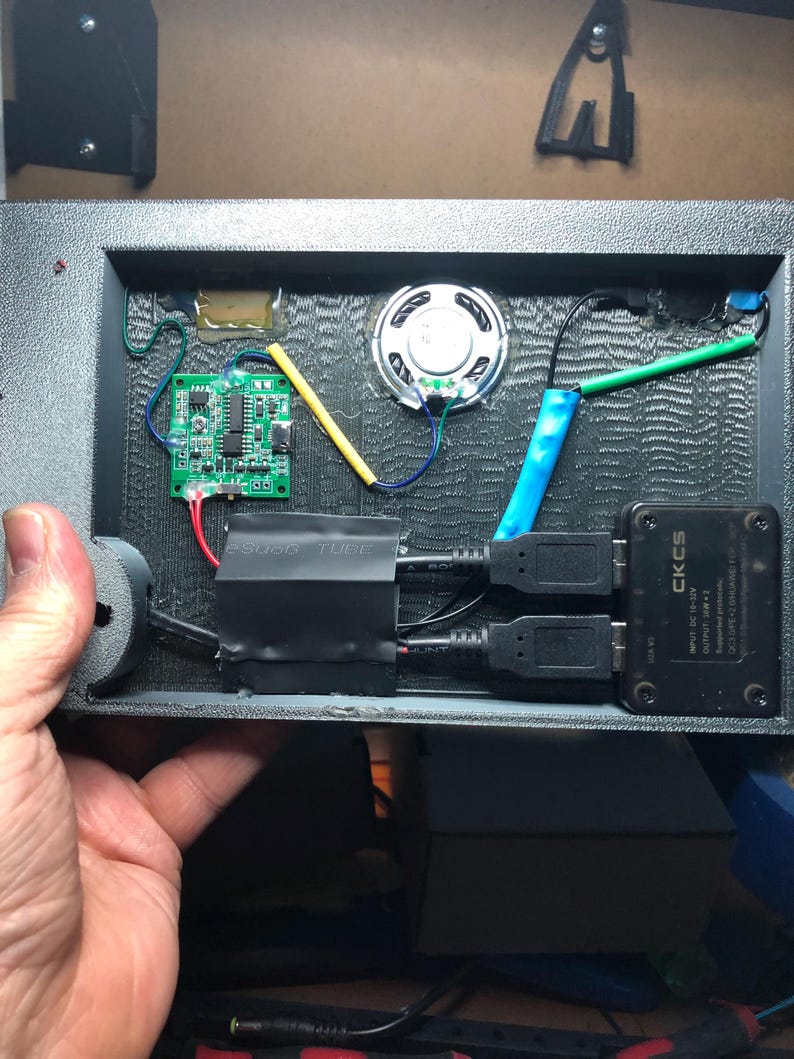 Może przedstawiać: Zbliżenie wnętrza czarnego urządzenia elektronicznego. Widoczne elementy to mały głośnik, zielona płytka drukowana oraz r&oacute;żne przewody i złącza. Urządzenie posiada r&oacute;wnież dwa porty USB i czarną skrzynkę z napisem "CKCS".