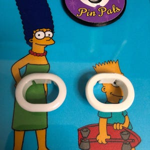 May include: Two white plastic rings with a blue background featuring cartoon characters from the Simpsons. The text "Pin Pals" is visible on a purple circle.