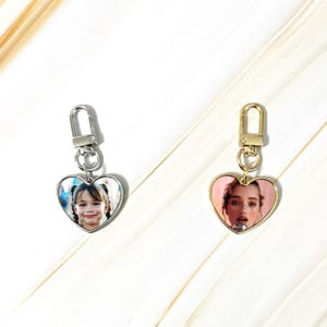 May include: Two heart-shaped keychains with photos. One keychain has a silver finish, the other gold. Both have a photo of a person inside the heart. The keychains have a clasp for attaching to keys or a bag.