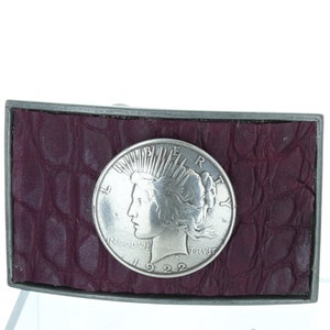 Puede incluir: Una hebilla de cinturón de plata con una moneda de 1922 Peace Dollar incrustada en el centro. La moneda está rodeada de un fondo de cuero burdeos con estampado de piel de cocodrilo.