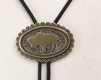 May include: A black bolo tie with a gold-toned oval pendant featuring a cutout silhouette of a buffalo. The pendant has a decorative border.