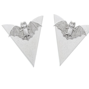 Może przedstawiać: Para srebrnych kolczyków w kształcie trójkąta z zawieszką w kształcie nietoperza wiszącą z każdego kolczyka.