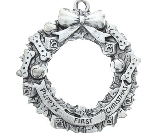 May include: A silver-toned Christmas ornament shaped like a wreath with a bow on top. The wreath is decorated with paw prints, bones, and bells. The text "Puppy's First Christmas" is inscribed on the wreath.