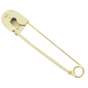Könnte beinhalten: Eine goldene Sicherheitsnadel mit einem abgerundeten Kopf und einem federbelasteten Verschluss.