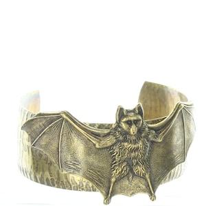 Può includere: Un bracciale a polsino color bronzo con un design dettagliato a pipistrello. Il pipistrello è centrato sul bracciale con le ali spiegate. Il bracciale ha una superficie strutturata e un design leggermente aperto.