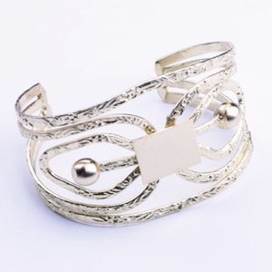 Könnte beinhalten: Silberarmband mit strukturiertem Design und zwei kleinen runden Akzenten. Das Armband ist offen und hat eine rechteckige Form in der Mitte.