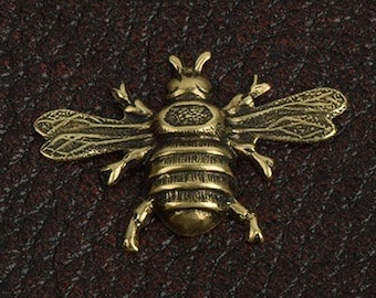 May include: A gold-coloured metal bee charm with detailed wings and a textured body. The bee is on a dark brown textured background.