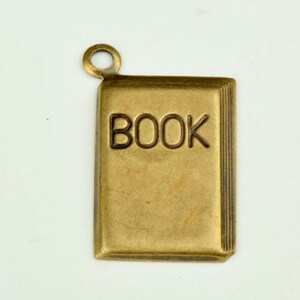 Puede incluir: Un amuleto de metal de color dorado con forma de libro con la palabra "BOOK" en relieve en la parte delantera.