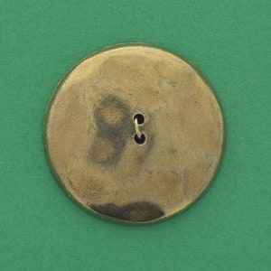 May include: A round, gold-colored button with a single hole in the center. The button is on a green background.