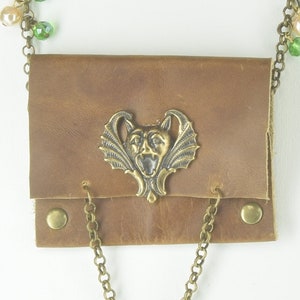 May include: A brown leather pouch with a brass chain and a decorative brass gargoyle pendant. The chain has green and cream beads.