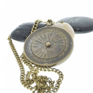 Puede incluir: Un collar de brújula de latón con una cadena y una cara de brújula circular con números romanos y marcas. El collar está apoyado sobre dos piedras lisas de color gris.