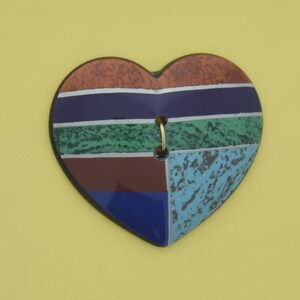 May include: A heart-shaped ceramic ornament with a geometric design in shades of blue, green, brown, and orange. The ornament has a gold ring in the centre.