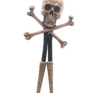 May include: A bronze skull and crossbones bolo tie with black cord. The skull is detailed with eye sockets, a nose, and a mouth. The bolo tie has two tassels at the bottom.