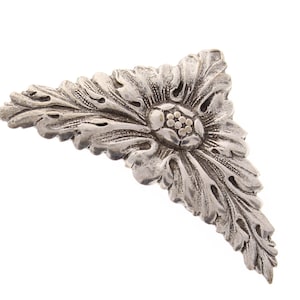 May include: A silver-toned, triangular-shaped decorative piece with a floral design. The piece has a raised, textured flower in the center with a cluster of small, raised dots.