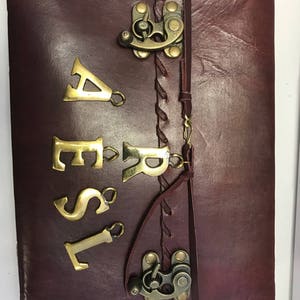 Puede incluir: Un diario de cuero burdeos con un cierre de latón y una inscripción de latón que dice "AESIR".