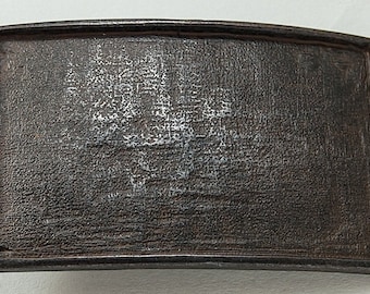May include: A rectangular, dark brown metal belt buckle with a slightly curved top edge. The surface has a textured appearance with a lighter, possibly corroded, design in the center. The buckle has a raised border.