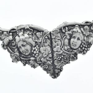 Könnte beinhalten: Eine silberfarbene Brosche aus Metall mit zwei weiblichen Gesichtern, umgeben von Weinreben und Blättern.